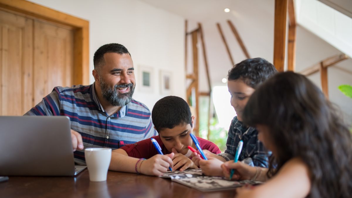 A parent working at a laptop at a table beside two children doing their own work