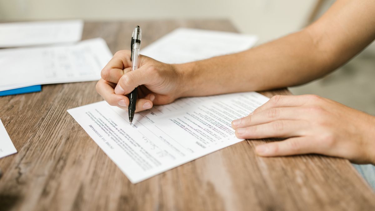A hand filling out a printed form with a pen on a wooden desk